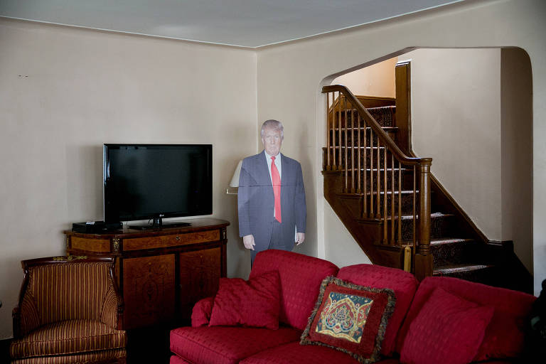 Um recorte de papelão do presidente Donald Trump em sua casa de infância no Queens, com uma televisão sobre um móvel de madeira, um sofá vermelho e uma escada lateral de acesso ao primeiro andar