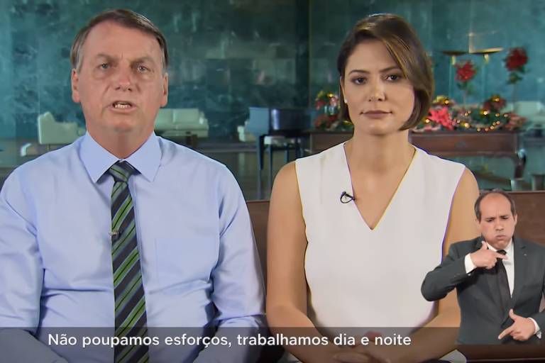 O presidente, de gravata e sem paletó, ao lado da primeira-dama, Michelle Bolsonaro, com uma roupa clara, sem mangas; na imagem também está presente um tradutor de libras