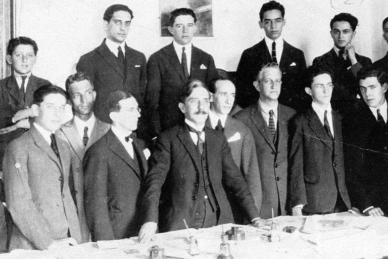 retrato em preto e branco de grupo de homens brancos vestindo terno posando para foto junto a uma mesa  