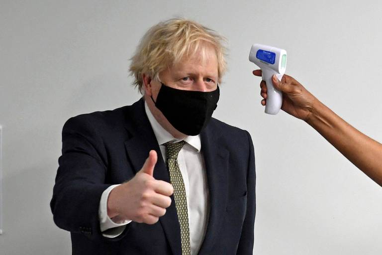 De máscara preta,paletó preto e camisa branca, Boris faz sinal de positivo com o polegar, enquanto se vê um braço e uma mão segurando um medidor de temperatura
