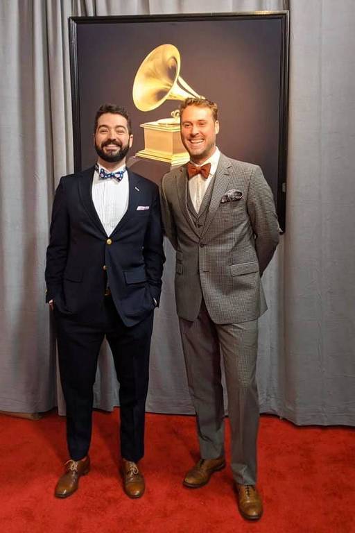 Dois homens de terno em tapete vermelho durante a cerimônia do Grammy 2020. Atrás deles, há uma imagem do troféu da premiação