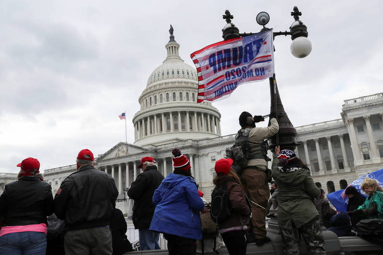 A imagem mostra um grupo de pessoas reunidas em frente ao edifício do Capitólio dos Estados Unidos. Algumas pessoas estão vestindo chapéus vermelhos e roupas de inverno. Uma pessoa está segurando uma bandeira que diz 'TRUMP 2020'. O céu está nublado e o edifício do Capitólio é claramente visível ao fundo.