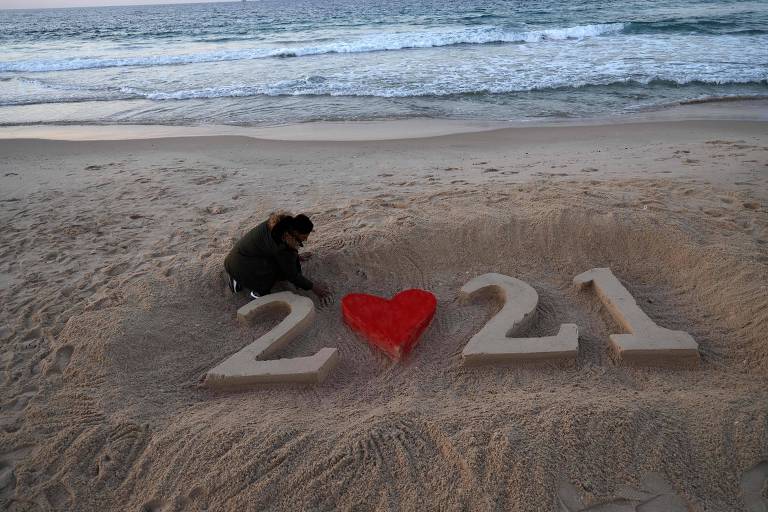 Mulher desenha em areia da praia numeral 2021, com um coração vermelho no lugar do 0
