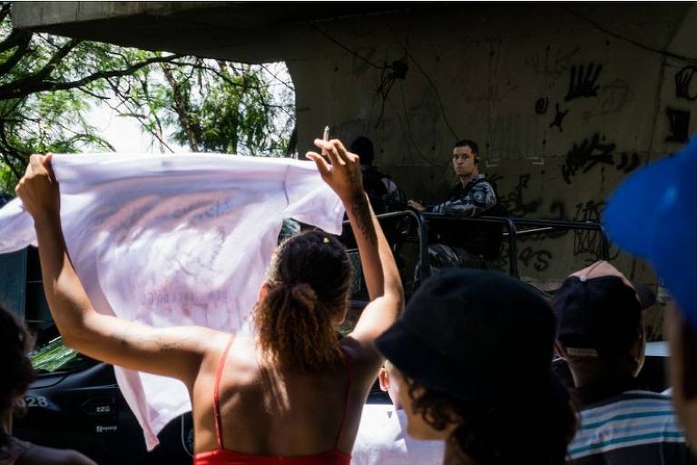 Grupo de manifestantes de costas para a câmera em frente a policiais; uma mulher segura uma camiseta branca e a ergue