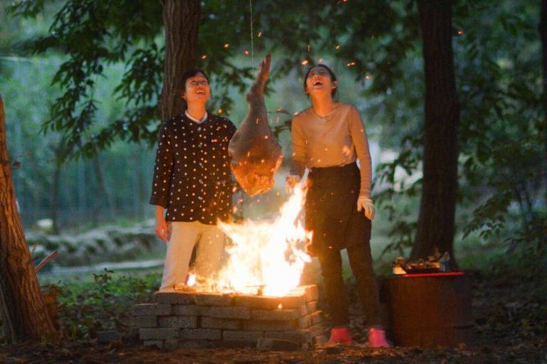 Duas mulheres estão atrás de uma fogueira no chão e tem um pernil pendurado em cima do fogo. O ambiente em que elas estão é uma floresta