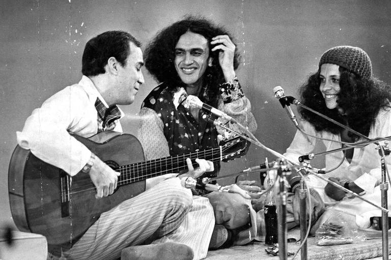 Dois homens e uma mulher num palco. Um deles toca violão e os outros sorriem