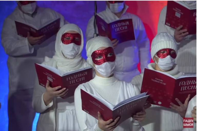 Cantores com roupas brancas e cabelos cobertos de branco e máscara vermelha sobre os olhos, segurando álbuns de partitura vermelhos