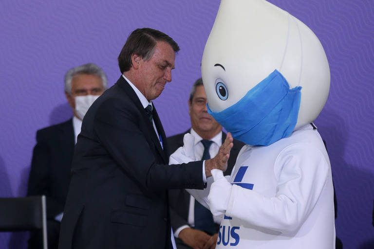 Bolsonaro, sem máscara, bate a mão no peito do Zé Gotinha, que usa uma máscara azul