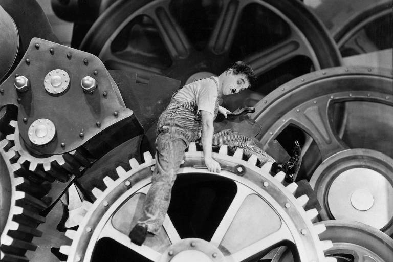 No filme em preto e branco, e personagem de Charlie Chaplin aparece sentado na engrenagem da máquina de uma fábrica