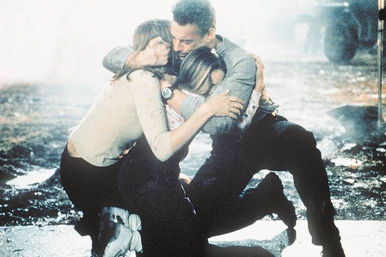 um homem agachado abraçando duas mulheres; explosão ao fundo