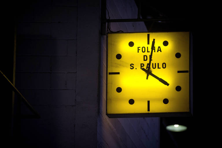 Relógio quadrado amarelo com bolinhas indicando os números e escrito Folha de S. Paulo