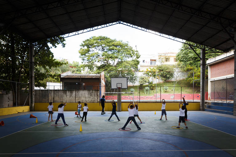 A imagem mostra uma quadra de esportes coberta, onde várias crianças estão participando de uma aula de educação física. Elas estão distribuídas em diferentes posições, algumas segurando bolas, enquanto outras estão em movimento. O ambiente é iluminado naturalmente, com árvores visíveis ao fundo e uma cesta de basquete ao centro da quadra. O piso é colorido, com círculos e cones de sinalização espalhados.