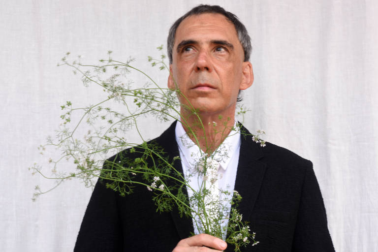 Vestindo paletó preto e camisa e gravata brancas, Arnaldo Antunes, moreno e grisalho, segura um ramo de coentro com flores