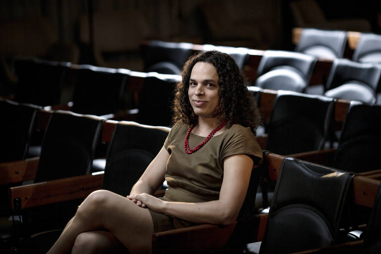 A escritora Amara Moira sentada em auditório. Ela está de vestido marrom e cabelo solto, abaixo dos ombros
