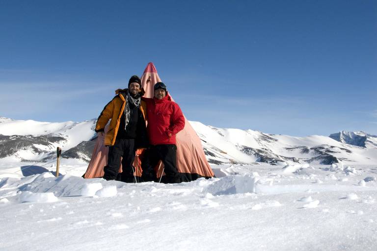 Dois homens com roupas de frio em frente a barraca no meio da neve e gelo