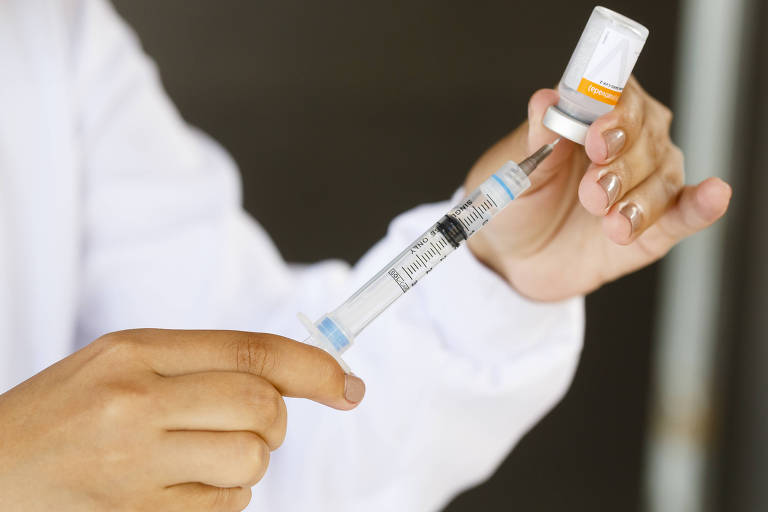 Imagem em close mostra uma pessoa segurando uma seringa e a espetando em uma ampola de vacina.