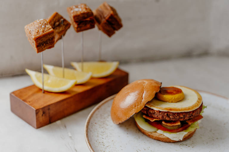 A imagem mostra um hambúrguer em um prato branco. O hambúrguer está montado com alface, tomate, cebola, picles e queijo, dentro de um pão de hambúrguer. Ao lado, há uma tábua de madeira com três espetos contendo pedaços de pancetta à base de proteína e fatias de limão.