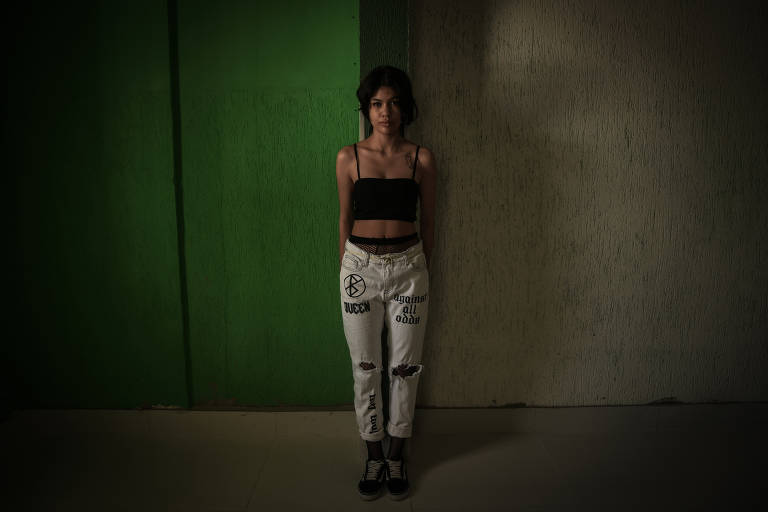 Uma jovem de cabelos pretos curtos vestida com calça jeans com rasgos, e top preto está de pé em frente a uma parede verde