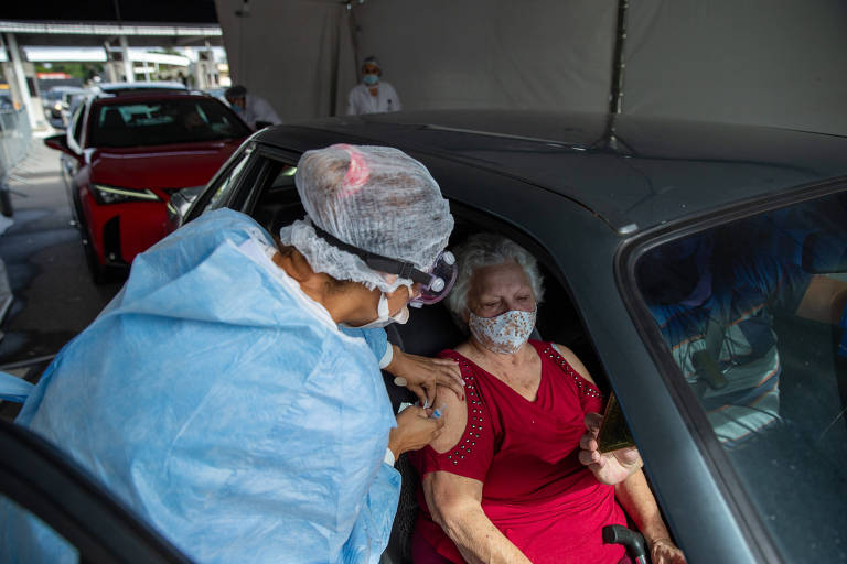 Enfermeira paramentada com equipamentos de segurança vacina idosa vestida com blusa vermelha. A idosa está sentada no banco do passageiro do carro. 