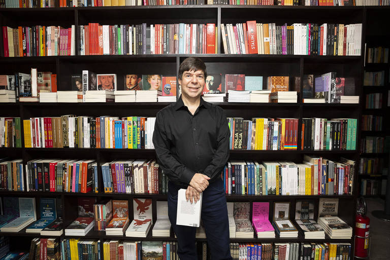 Homem branco usa terno preto e sorri em frente a estante de livros