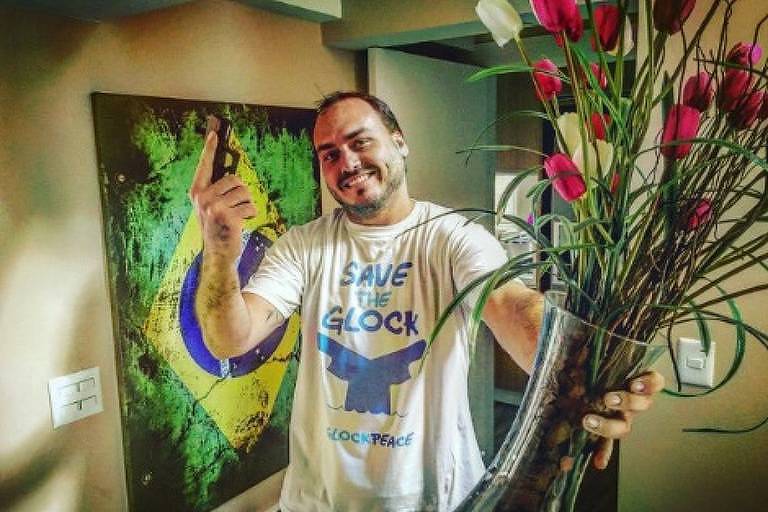 -Carlos Bolsonaro com uma arma na mão direita e um vaso de flores na esquerda; ele sorri para a câmera e usa uma camiseta que diz "save the Glock" (salve a Glock, sendo Glock uma fabricante de armamentos); ao fundo há um quadro reproduzindo a bandeira do Brasil, mas orientada na vertical