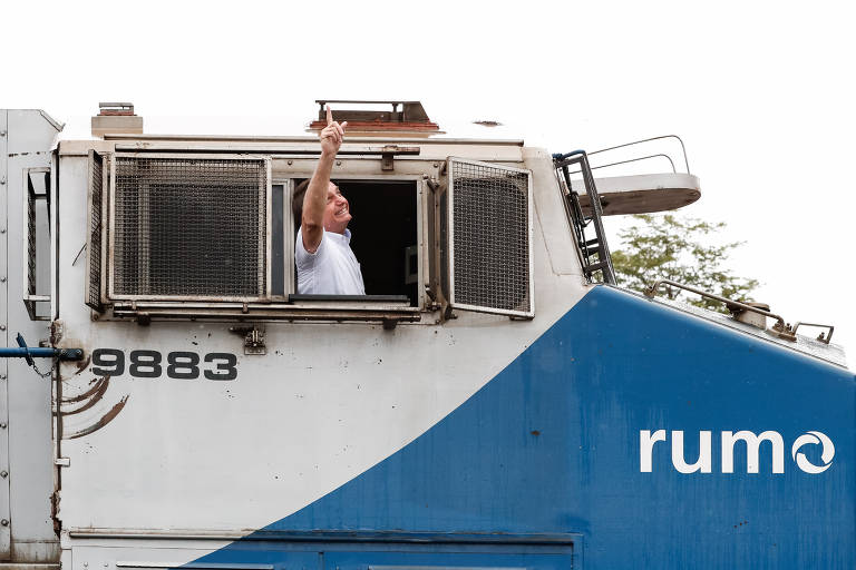 Sentado dentro do vagão de frente de um trem em que se lê a palavra "rumo", o presidente Jair Bolsonaro, vestindo camisa branca de manga curta, olha e aponta em direção ao céu