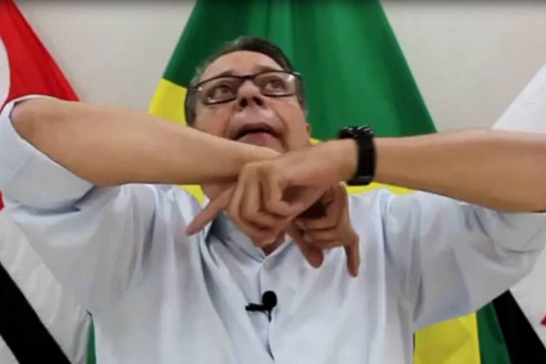 homem de camisa de manga comprida gesticula com as duas mãos; ao fundo, bandeiras de São Paulo, do Brasil e de Pirassununga