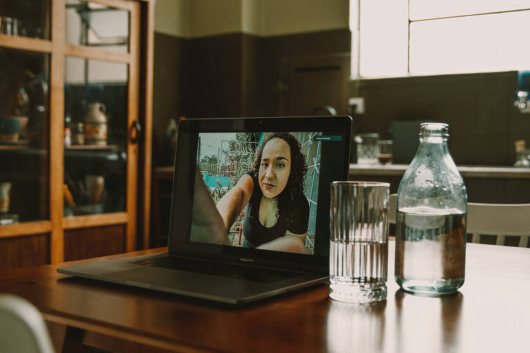 Foto feita a distância mostra, na tela de um computador, uma jovem parda, de cabelos cacheados; na mesa onde está o laptop há uma garrafa e um copo de água