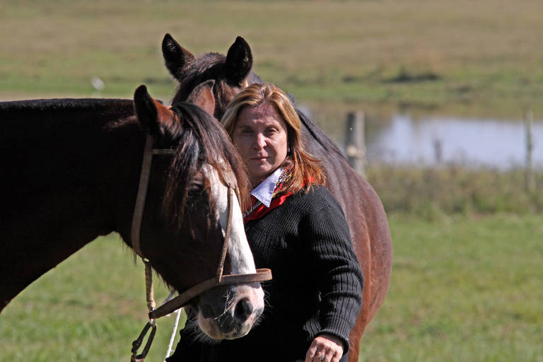 Mulher de cabelos castanhos calros lisos, paletó azul marinho e camisa branca, com dois cavalos