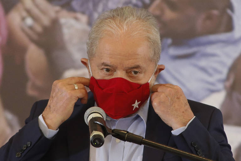Imagem em primeiro plano mostra Lula, de social, de frente para um microfone e colocando uma máscara vermelha