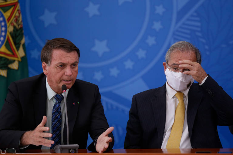 Dois homens brancos estão sentados; um está discursando enquanto o outro, que usa máscara contra a Covid-19, leva as mãos ao rosto