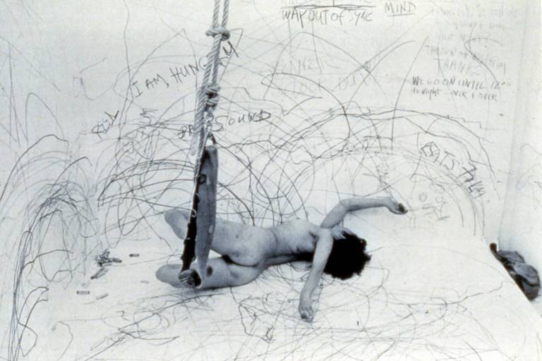 A imagem em preto e branco mostra uma mulher nua deitada em um cenário completamente branco, de paredes e chão rabiscados 