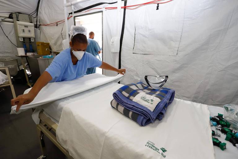 Funcionário com roupa azul e mascara dobra lençol de maca em tenda de hospital