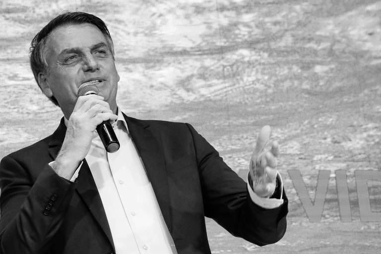 Presidente Jair Bolsonaro, do lado esquedo da foto, fala ao microfone vestindo camisa e paletó. Ele aparece apenas da cintura para cima. Com a mão esquerda, gesticula. O fundo da foto é azul e é possível ver parte do nome da Igreja escrito: "vida"