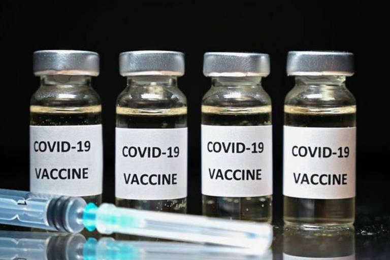 Quatro frascos de vacina contra a Covid-19 com uma seringa à frente, em fundo preto