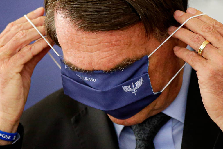 O presidente Jair Bolsonaro tenta colocar máscara com brasão da FAB durante anúncio de investimentos para o programa Águas Brasileiras, no Palácio do Planalto, em Brasília