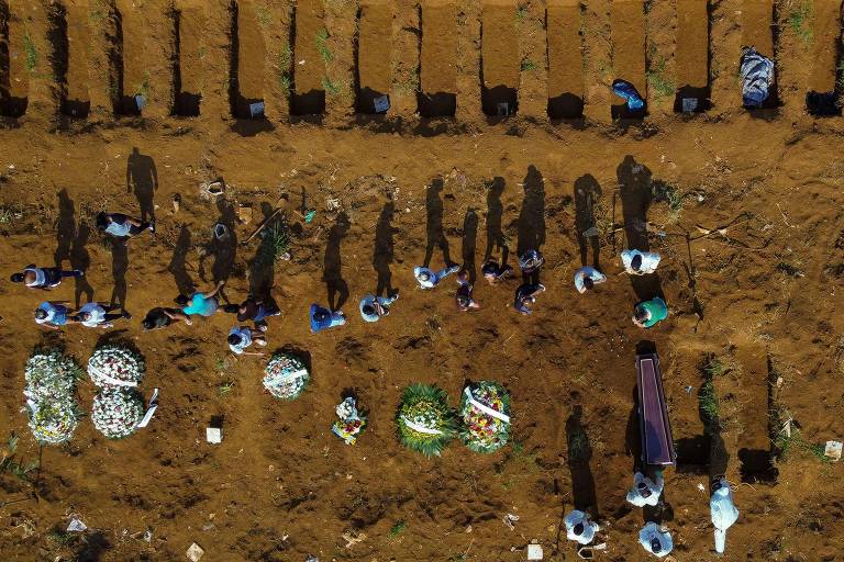 Vista aérea de duas fileiras de covas em parte fechadas e cobertas de flores. No meio, há uma fileira de pessoas acompanhando um enterro.