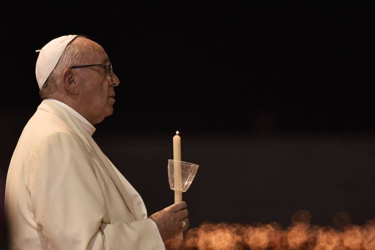 A imagem mostra um homem idoso vestindo trajes clericais brancos e um solidéu branco， segurando uma vela acesa com um suporte transparente. O fundo está escuro， com luzes difusas ao longe， sugerindo um ambiente religioso ou cerimonial， possivelmente uma vigília ou celebração noturna. A expressão do homem é séria e contemplativa， e ele parece concentrado no momento.