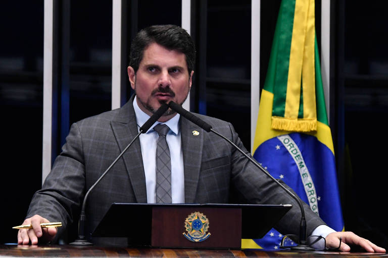 Homem branco, veste terno e gravata, discursa com as mãos apoiadas na tribuna, do seu lado esquerdo vê-se a bandeira do Brasil