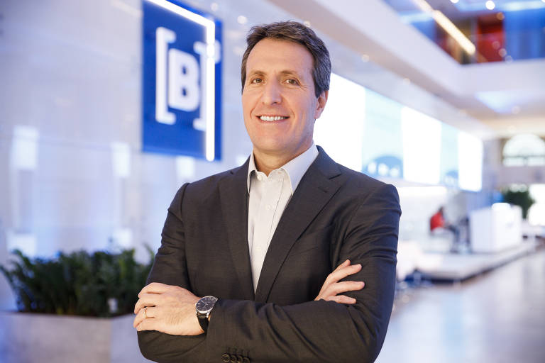 Um homem sorridente está posando em um ambiente corporativo moderno. Ele está vestido com um terno escuro e uma camisa clara， com os braços cruzados. Ao fundo， há uma decoração minimalista e uma grande placa com o escrito B3.