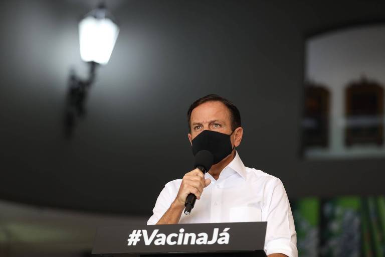 João Doria está de máscara preta em frente a púlpito com a inscrição "#VacinaJá", falando ao microfone