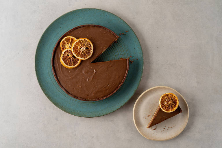 Em um prato azul, tem uma torta com base de cacau, recheio de amêndoas e cobertura de chocolate com laranja. Em um prato menor e branco ao lado, tem uma fatia dessa mesma torta.