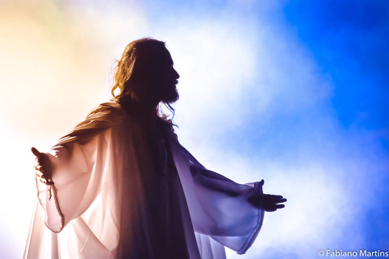 A imagem mostra a silhueta de uma figura religiosa com cabelos longos e vestes brancas, de braços abertos, contra um fundo de fumaça colorida em tons de azul e amarelo. A iluminação destaca a forma da figura, criando um efeito dramático.