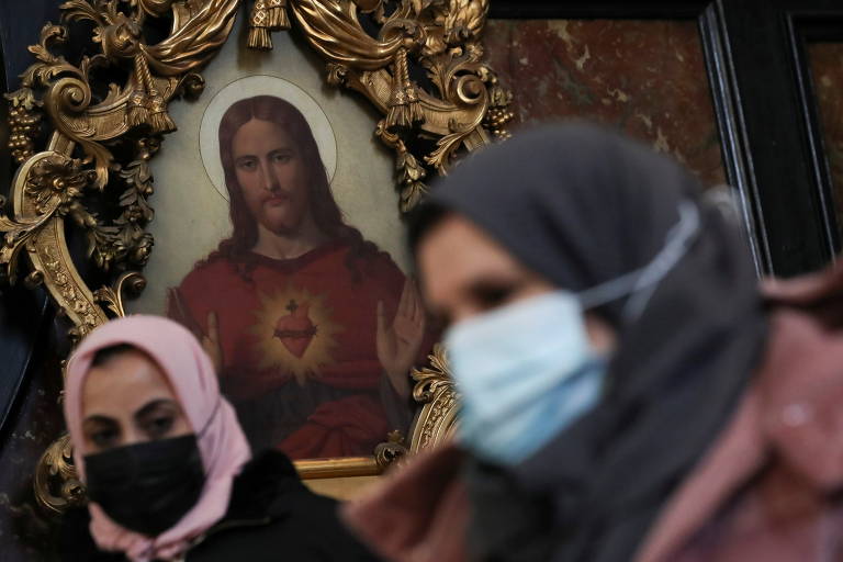Duas mulheres de máscaras e lenço cobrindo o cabelo, com uma pintura de Jesus Cristo ao fundo