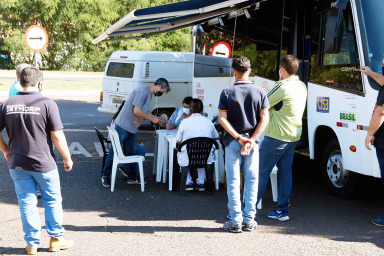Imagem mostra homens em pé, ao lado de trailer, enquanto aguardam atendimento em mesas instaladas em barreira sanitária