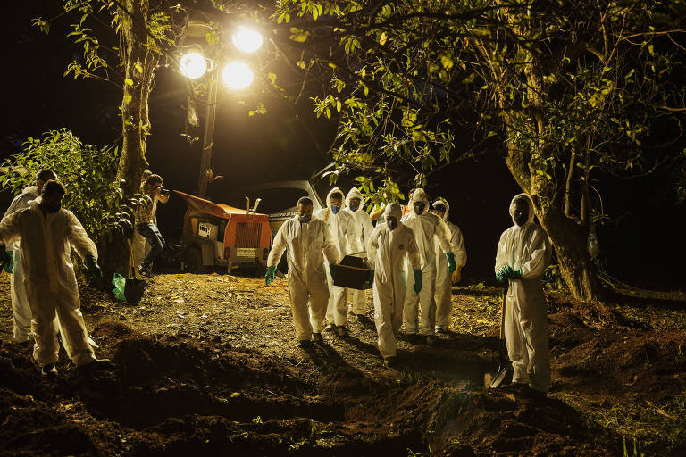 Vários homens com os corpos totalmente cobertos com roupas brancas carregam caixão para sepultamento em área verde à noite