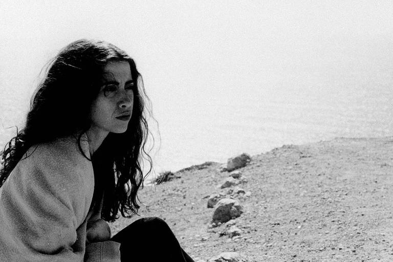 Uma jovem com cabelo longo e ondulado está sentada em uma superfície rochosa à beira do mar. Ela olha para o horizonte, com uma expressão pensativa. A imagem é em preto e branco. O mar é visível ao fundo, com uma linha do horizonte suave.