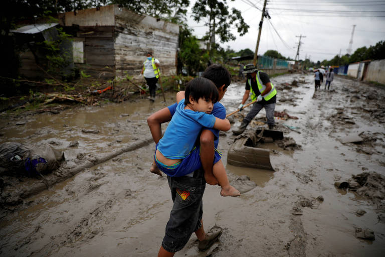 Menino carrega seu irmão em rua coberta de lama depois da passagem dos furacões Eta e Iota em La Lima (Honduras)