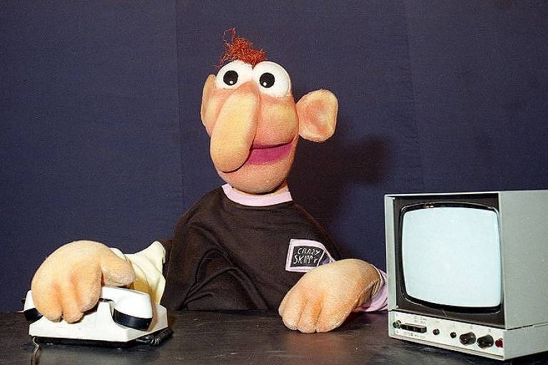 Boneco narigudo, de camiseta marrom, ao lado de uma TV e um telefone de brinquedo