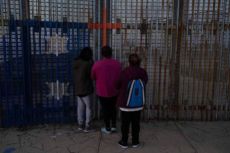 Migrantes olham através de cerca na fronteira dos EUA com o México, em Tijuana
